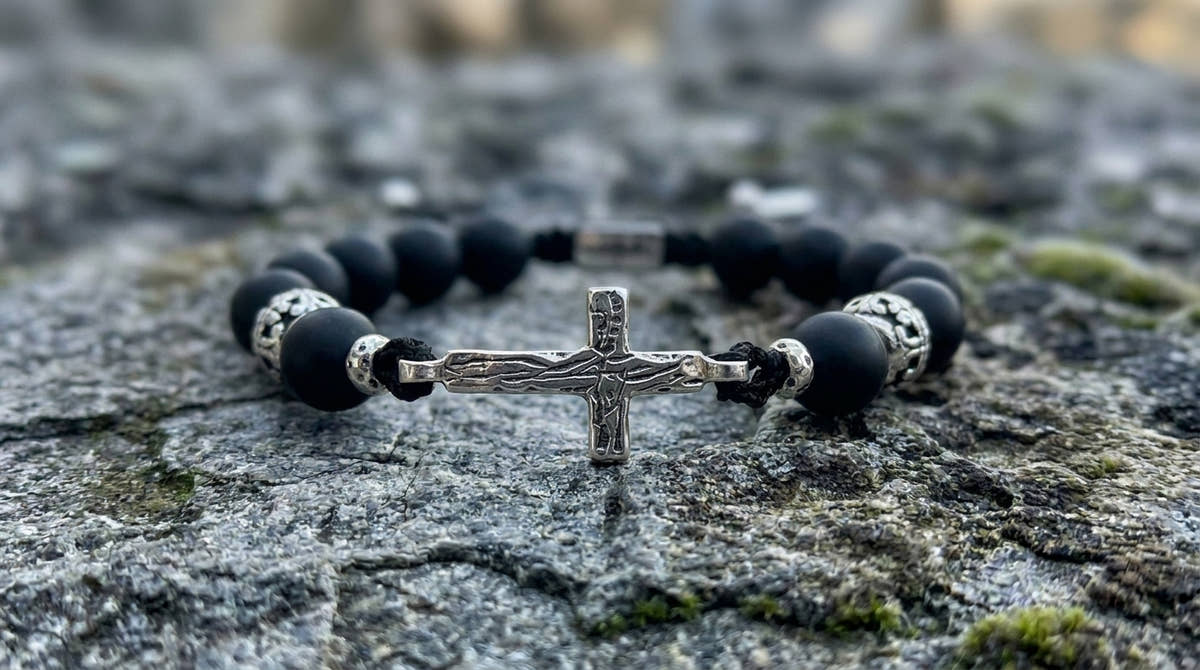 A beaded bracelet with matte black round beads, silver-toned spacer details, and a central silver crucifix charm, resting on a textured gray stone surface.
