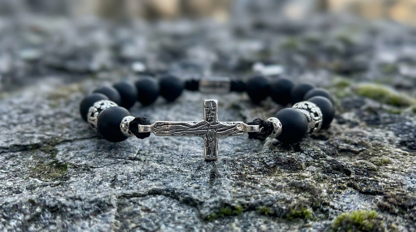 A beaded bracelet with matte black round beads, silver-toned spacer details, and a central silver crucifix charm, resting on a textured gray stone surface.