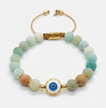 A beaded stretch bracelet with light blue, mint green, peach, and cream beads, accented by gold spacer beads and a central gold-plated evil eye charm. The bracelet is completed with a tan cord and a gold clasp engraved with 'cosmo'.